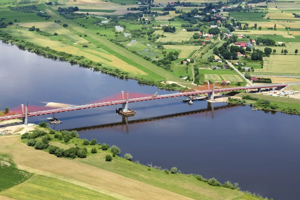 Przeprawa w Kwidzynie to most sprężony typu extradosed. Most na dużej rzece otoczony zielonymi polami i budynkami w oddali, ukazując spokojny pejzaż wiejski.