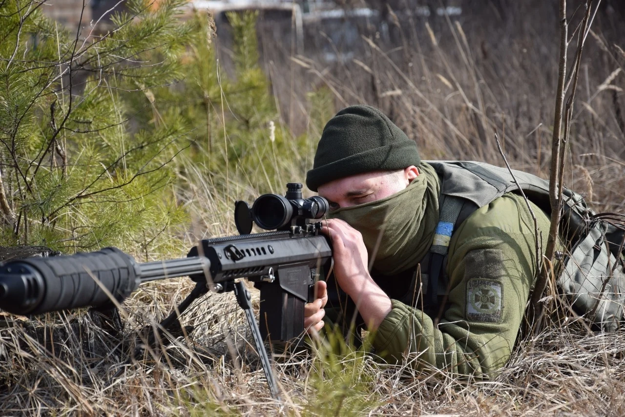 Żołnierz w stroju maskującym leży w trawie i celuje z karabinu snajperskiego. Otoczony jest roślinnością, co sugeruje, że znajduje się w terenie szkoleniowym lub polowym.