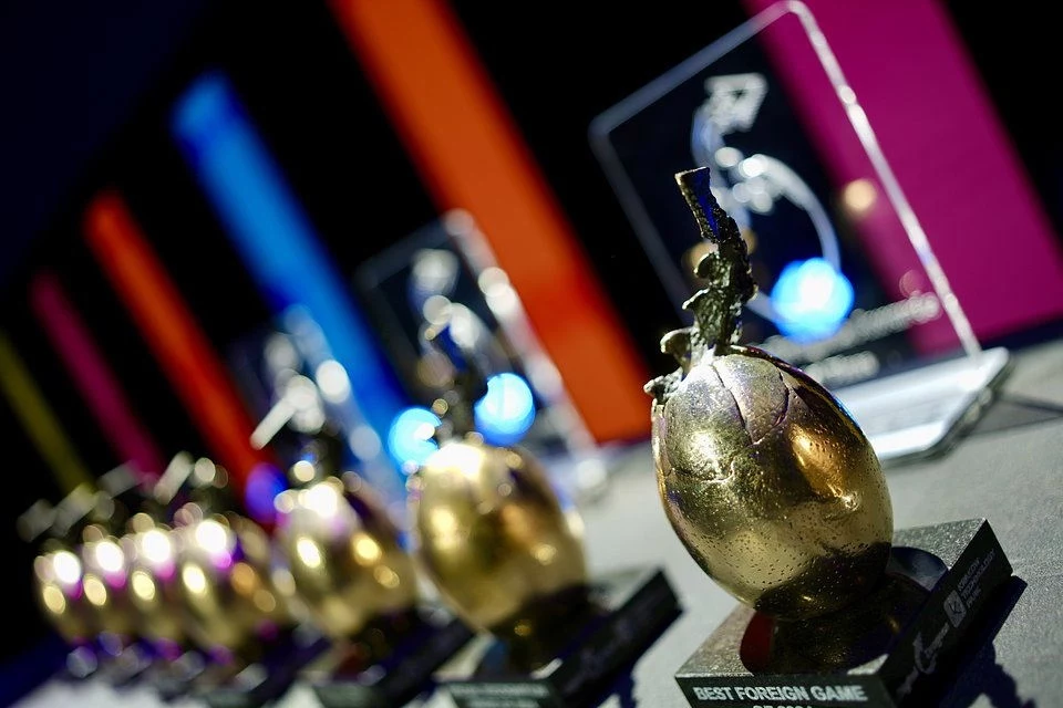 Trofea o charakterystycznym kształcie z napisem 'BEST FOREIGN GAME', ustawione na stole, błyszczące w mocnym oświetleniu. W tle znajdują się kolorowe, rozmyte światła tworzące dynamiczną atmosferę imprezy lub gali.