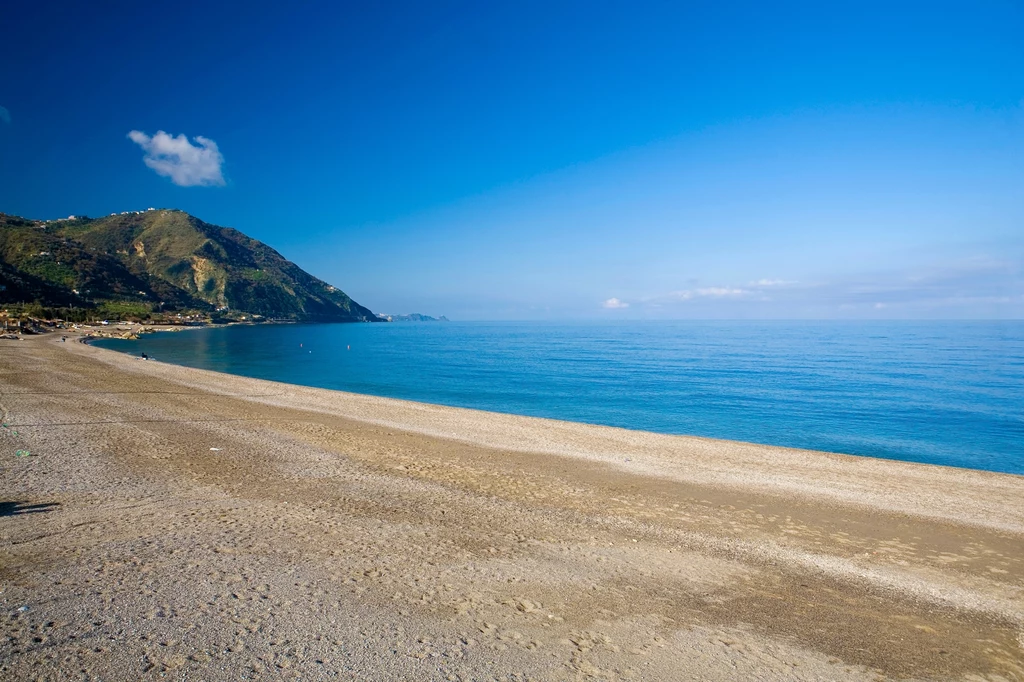 Plaże na Sycylii zachwycają błękitną, lazurową wodą i wszechobecnym spokojem Szeroka piaszczysta plaża u podnóża zielonych wzgórz, otulona spokojnymi wodami morza pod błękitnym niebem, które dodatkowo zdobią niewielkie chmury.