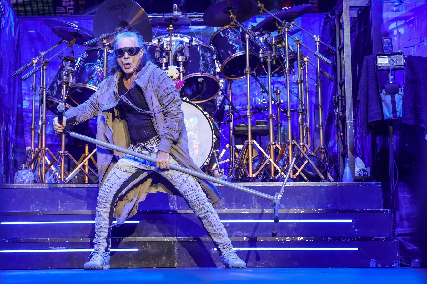 Bruce Dickinson (Iron Maiden) w akcji Mężczyzna w okularach przeciwsłonecznych i stroju sceniczny z długim płaszczem trzymający mikrofon na tle dużego zestawu perkusyjnego, scena oświetlona niebieskim światłem.