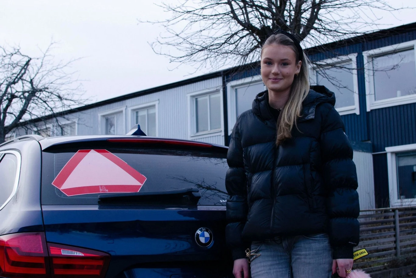 Młoda kobieta w kurtce puchowej stoi obok ciemnego BMW z dużym oznaczeniem na tylnej szybie. W tle widać jasny budynek i drzewa bez liści.
