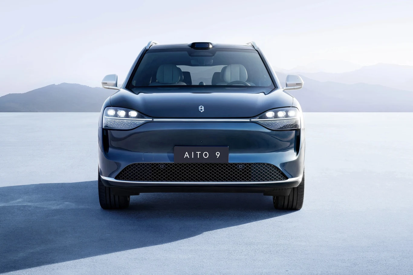 Elektryczne auto marki Aito zaparkowane na otwartej przestrzeni z górskim tłem. Przód pojazdu jest widoczny, z nowoczesnym designem i charakterystycznymi reflektorami. Jasne, minimalistyczne otoczenie podkreśla elegancki wygląd samochodu.