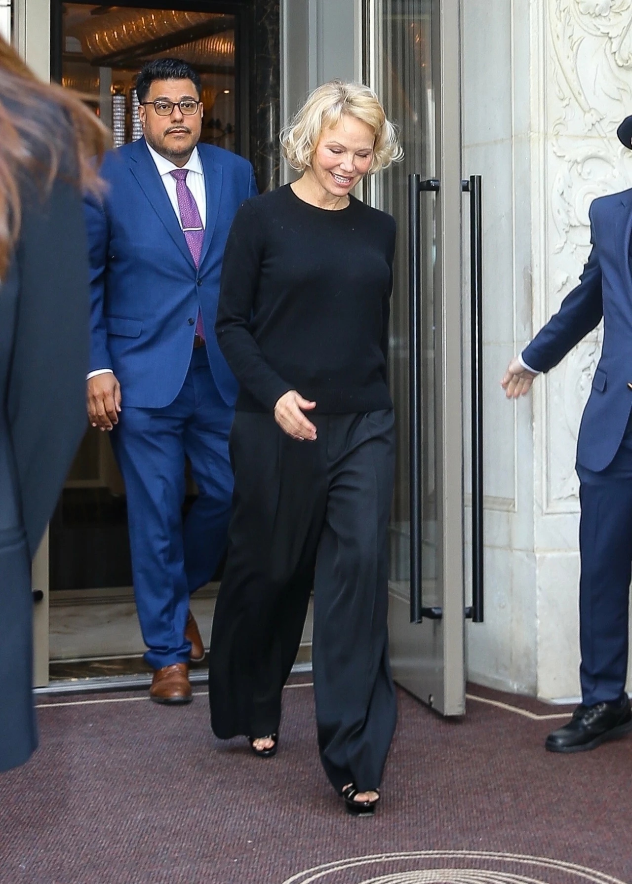 Pamela Anderson pożegnała swoje długie blond włosy Kobieta opuszczająca budynek przez szklane drzwi, ubrana w czarne spodnie i bluzkę. Towarzyszą jej dwaj elegancko ubrani mężczyźni w garniturach. W tle widoczna jest bogato zdobiona część wnętrza budynku.