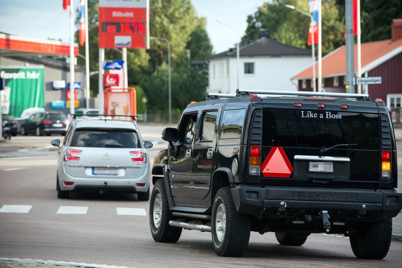 Czarny SUV z napisem 'Like a Boss' na tylnej szybie jedzie ulicą w małym mieście. Widoczne są również inne samochody, stacja benzynowa oraz domy w tle.