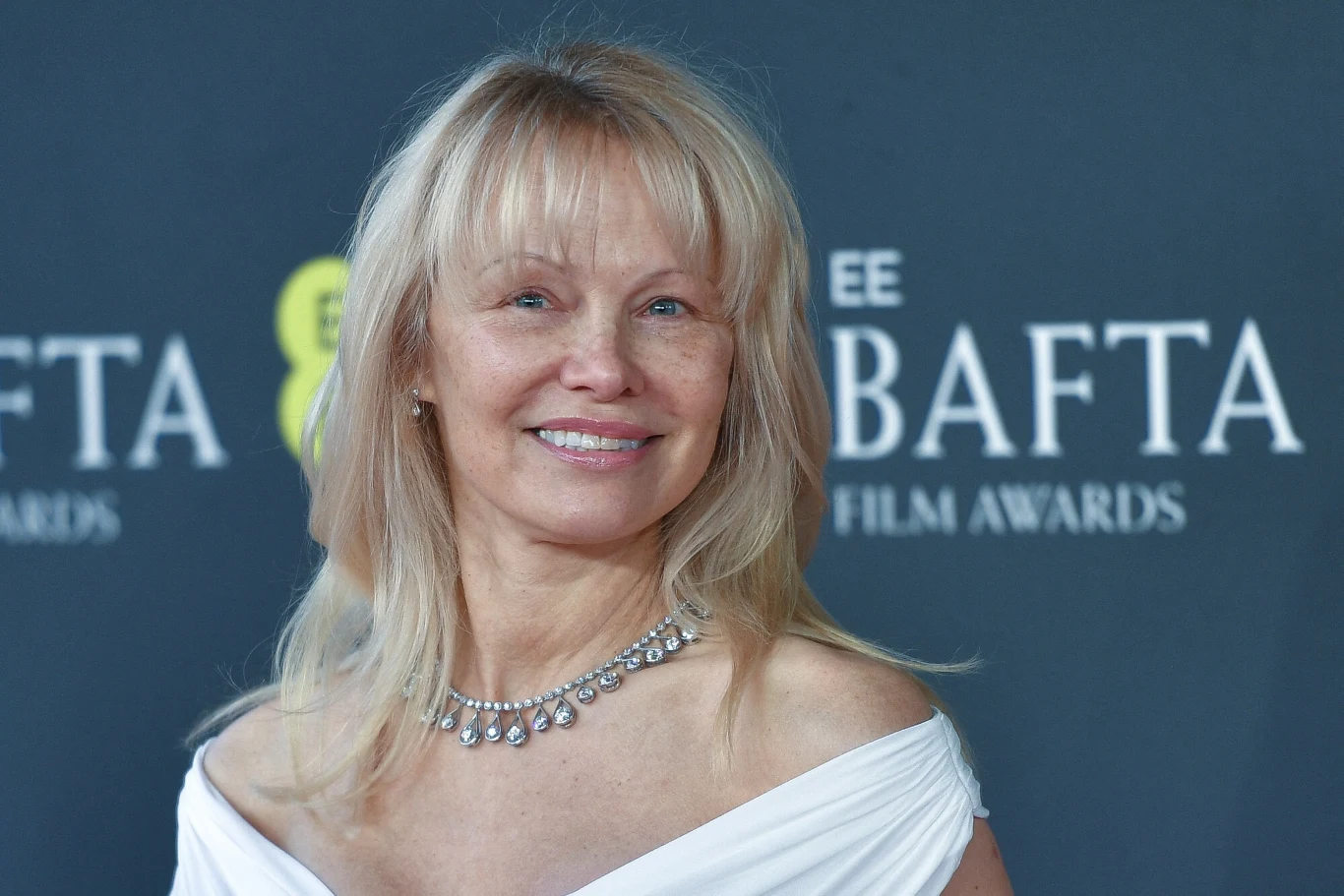 Pamela Anderson wciąż zaskakuje swoich fanów Kobieta o długich blond włosach i bez makijażu pozuje na ściance w białej sukni i srebrnej kolii na szyi