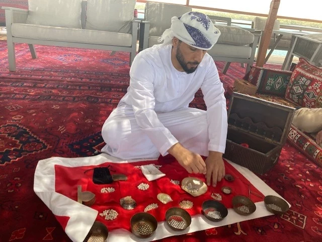 Suwaidi Pearls - pływająca platforma po brzegi wypełniona historią poławiaczy pereł w Ras al-Khaimah Mężczyzna w tradycyjnym stroju siedzi na dywanie, przeglądając metalowe miski z przyprawami lub ziołami, które leżą na białej tkaninie z czerwonymi akcentami.