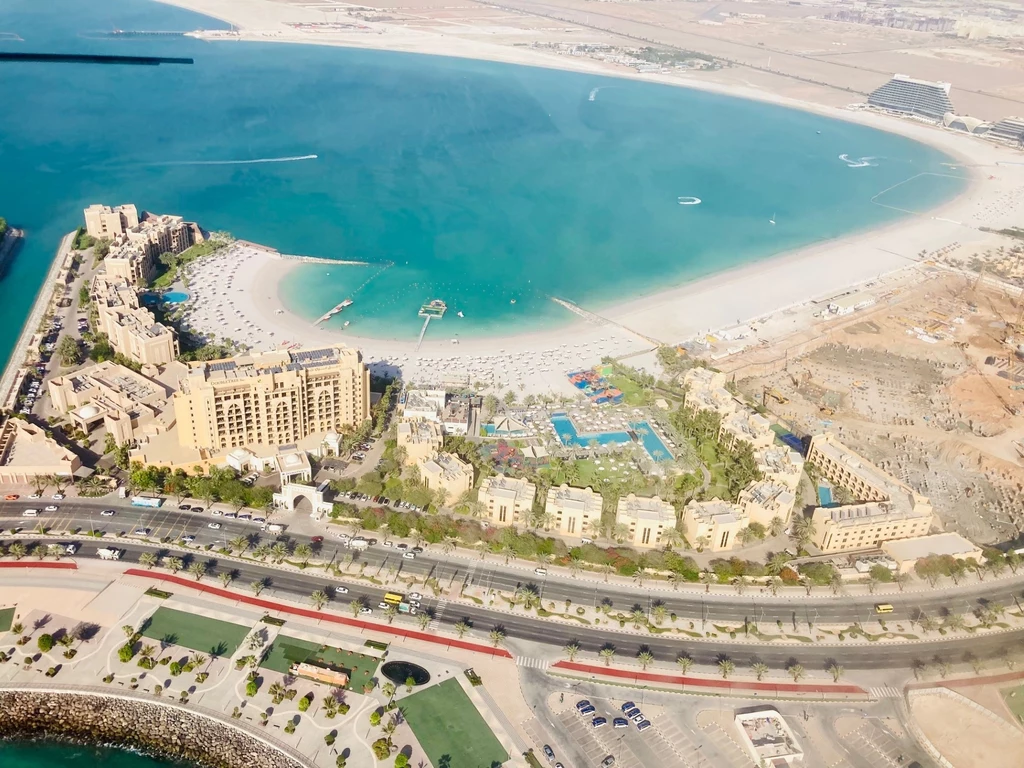 Kompleksy luksusowych hoteli w Ras al-Khaimah robią wrażenie, zwłaszcza z lotu ptaka Widok z lotu ptaka na nadmorski kompleks hotelowy z luksusowymi budynkami i basenami. Wokół rozciąga się biała, piaszczysta plaża oraz błękitne wody zatoki. W tle widać niewielkie łodzie i pustynny krajobraz.