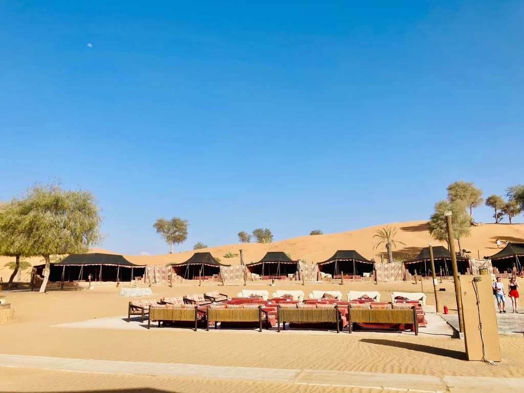 Bassata Camp Village w Ras al-Khaimah Pustynny krajobraz z szeregiem czarnych namiotów, otoczony palmami i suchymi drzewami. Na pierwszym planie znajdują się kanapy z czerwonymi poduszkami, a w tle widać złote wydmy pod błękitnym niebem.