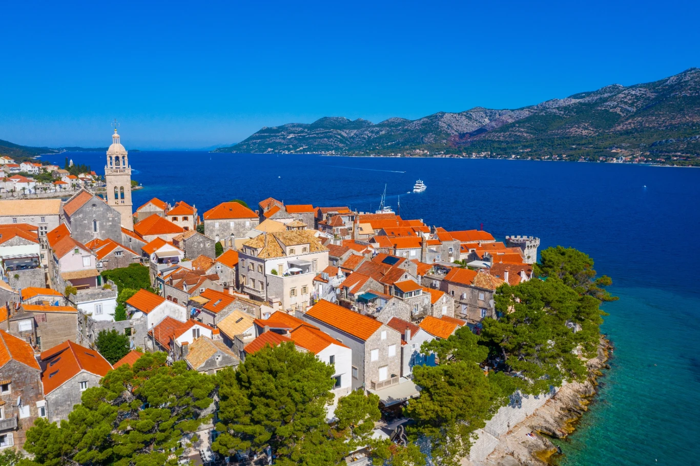 Aerial view of a coastal town with red-tiled roofs, bordered by the blue sea and mountains on the horizon.
