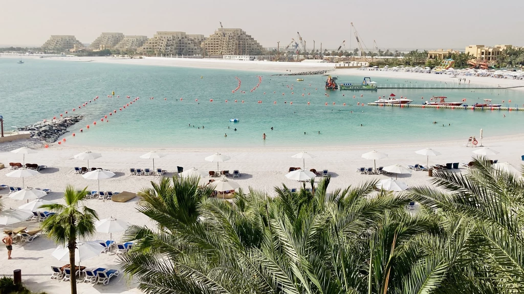 Cudowne plaże w Ras al-Khaimah wyglądają niczym z pocztówki Plaża z widokiem na spokojne morze, na tle którego znajdują się luksusowe budynki i żurawie budowlane. Białe leżaki i parasole ustawione w rzędach wzdłuż brzegu, otoczone palmami.
