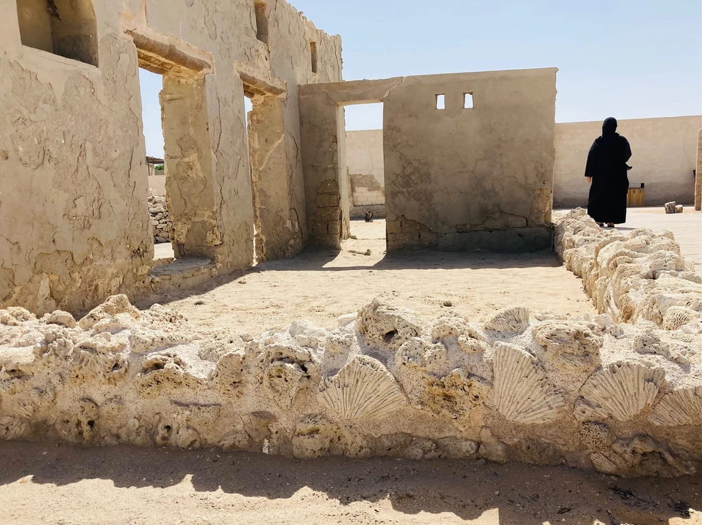 Al Jazeera Al hamra - jedyna zachowana wioska poławiaczy pereł Ruiny starego budynku z murami z kamieni i muszli, osoba w czarnej szacie stojąca w oddali, wnętrze oświetlone słońcem.