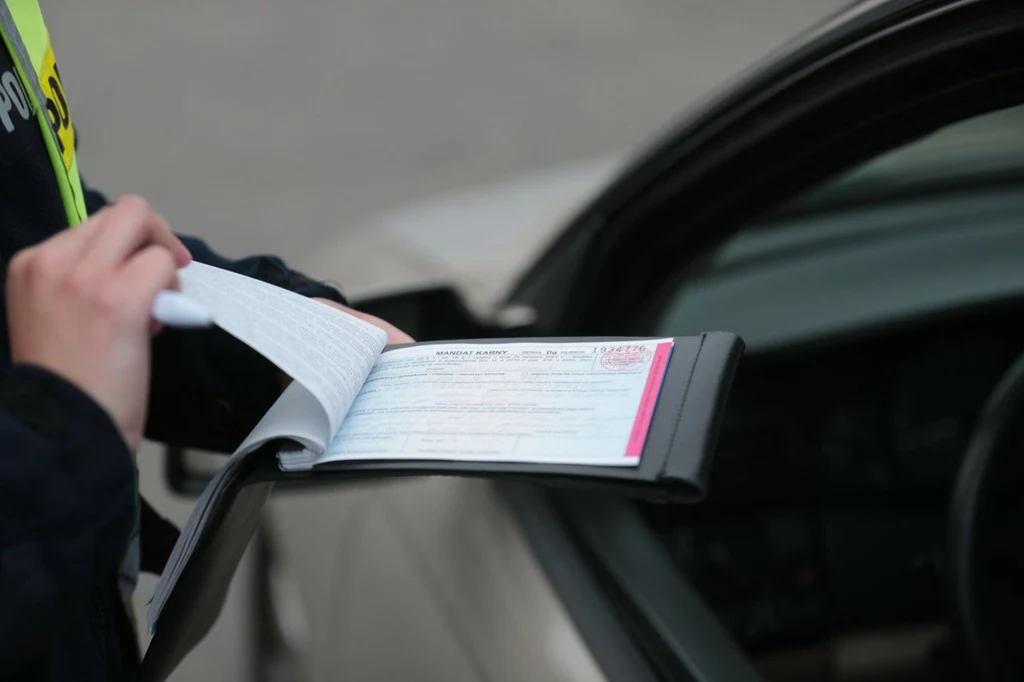 Policja wystawiła mandaty uczestnikom zdarzenia. Fragment samochodu z otwartymi drzwiami i osoba wypisująca mandat na bloczku, trzymająca w ręku długopis.