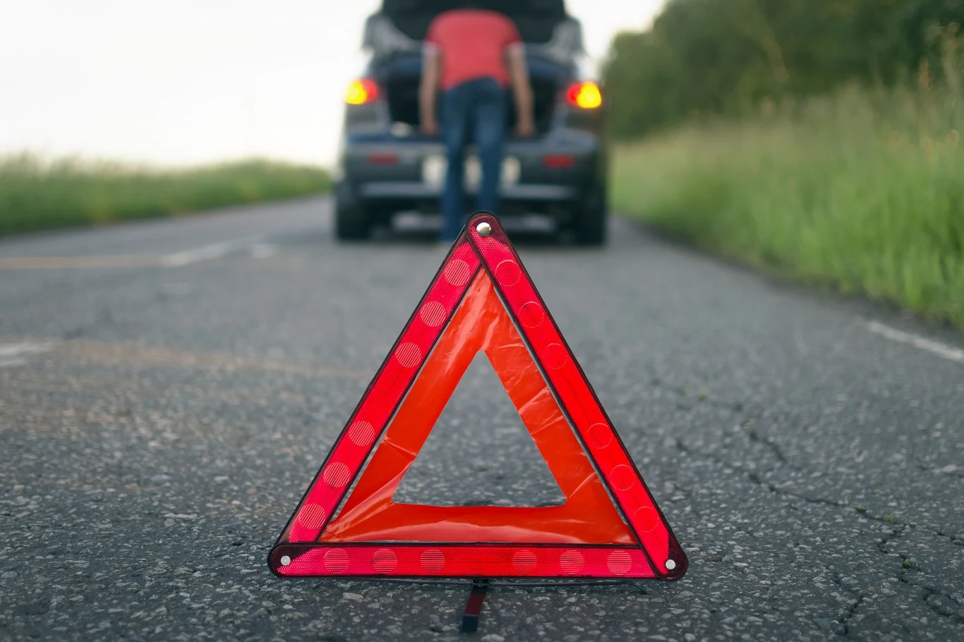 W jakich sytuacjach należy używać świateł awaryjnych? Trójkąt ostrzegawczy ustawiony na środku drogi, w tle widoczny samochód z otwartą maską i osoba pochylająca się nad silnikiem. Droga otoczona zielenią, z przewagą prostych linii i naturalnego światła.