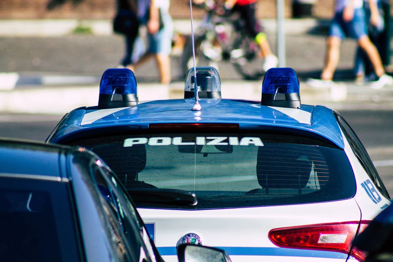 Policyjny samochód oznaczony napisem 'Polizia', na dachu widoczne są niebieskie światła sygnalizacyjne. W tle widać pieszych przekraczających ulicę.