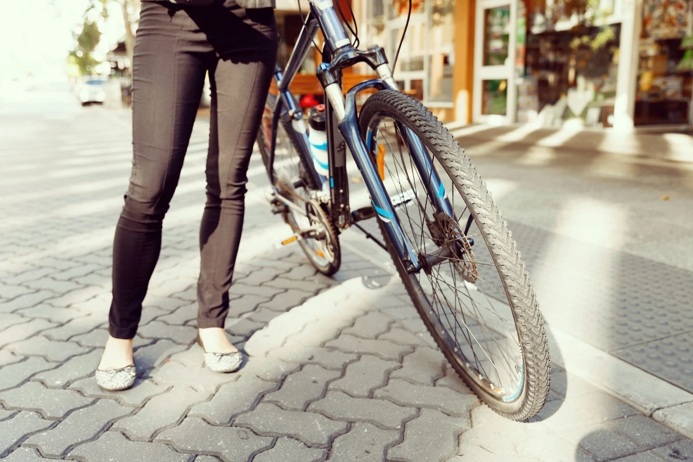 Osoba stojąca obok roweru na chodniku, widoczna od pasa w dół. W tle można dostrzec elementy miejskiej zabudowy.