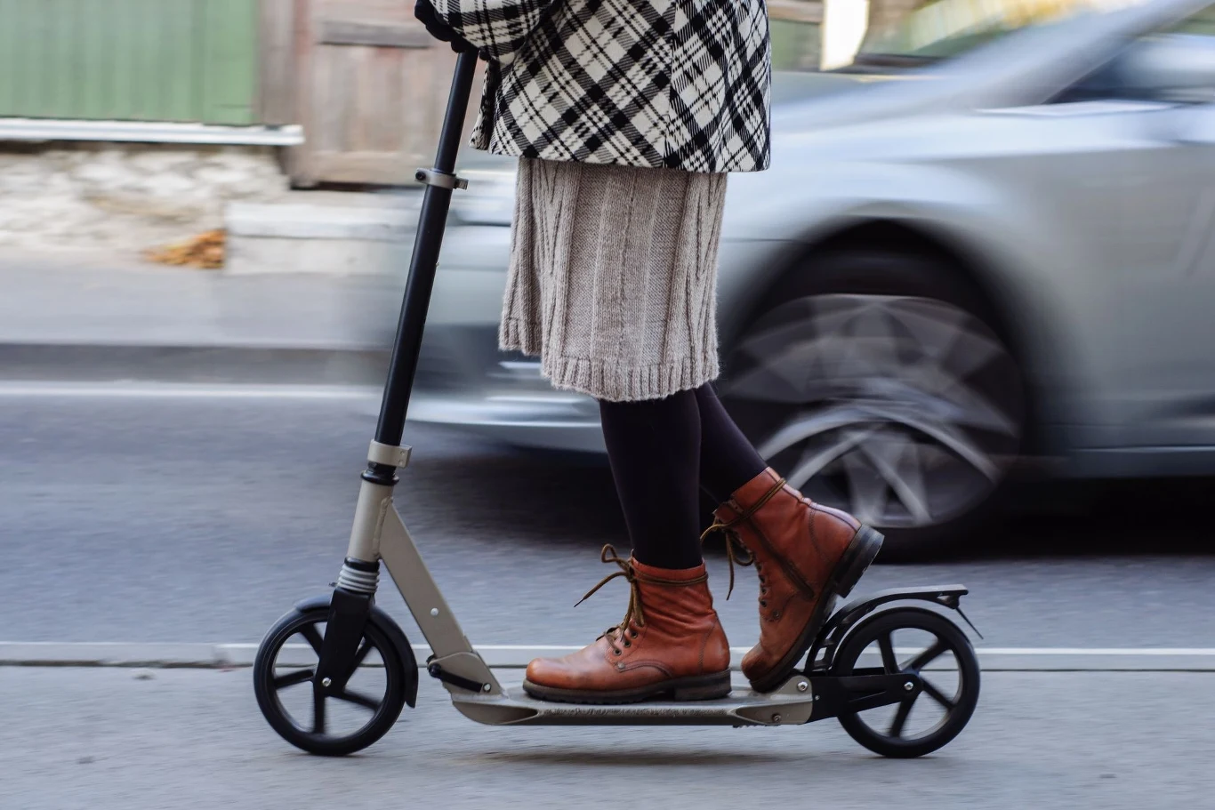 Osoba jedzie na hulajnodze po chodniku, ubrana w płaszcz w kratę, spódnicę i brązowe buty. Obok mija ją samochód w ruchu.