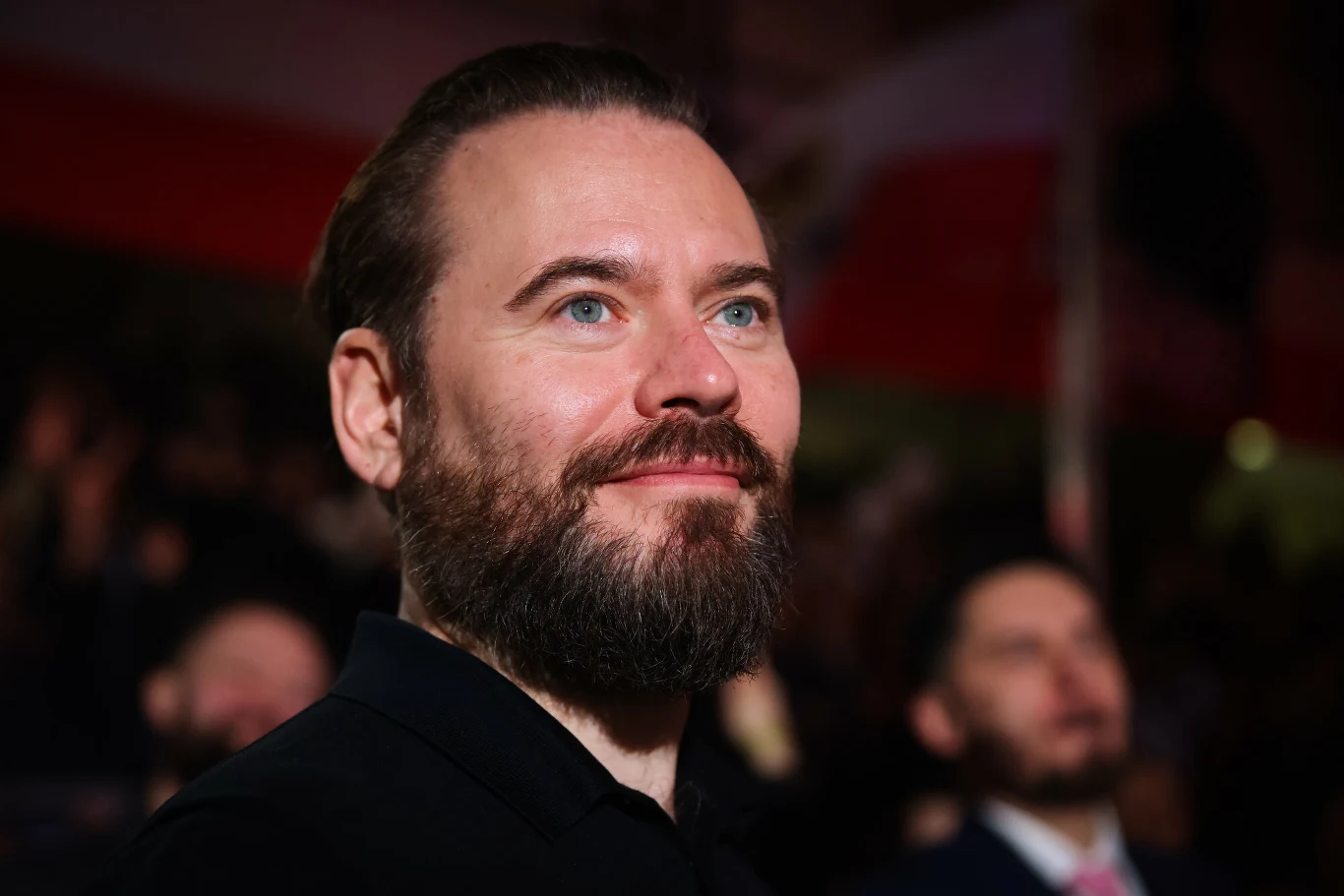 Mężczyzna z brodą patrzy w górę z delikatnym uśmiechem. Tło rozmazane, widać innych ludzi i światła w odległości.