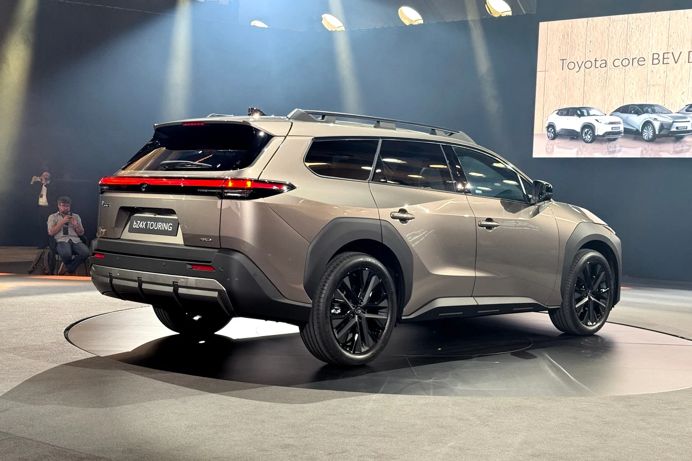 Nowoczesny samochód SUV zaprezentowany w sali wystawienniczej. Posiada elegancką, futurystyczną sylwetkę i ciemne wykończenia detali zewnętrznych. W tle ekran z informacją o innych samochodach marki Toyota.