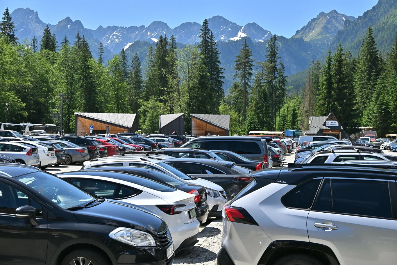 Parkingi na Łysej Polanie stanowią ostatni punkt legalnej podróży samochodem. Parking pełen samochodów, otoczony górami i lasem. W tle widoczne są szczyty górskie z ośnieżonymi wierzchołkami. Na pierwszym planie znajduje się wiele zaparkowanych aut. Sceneria sugeruje górzyste miejsce turystyczne, popularne wśród odwiedzających.