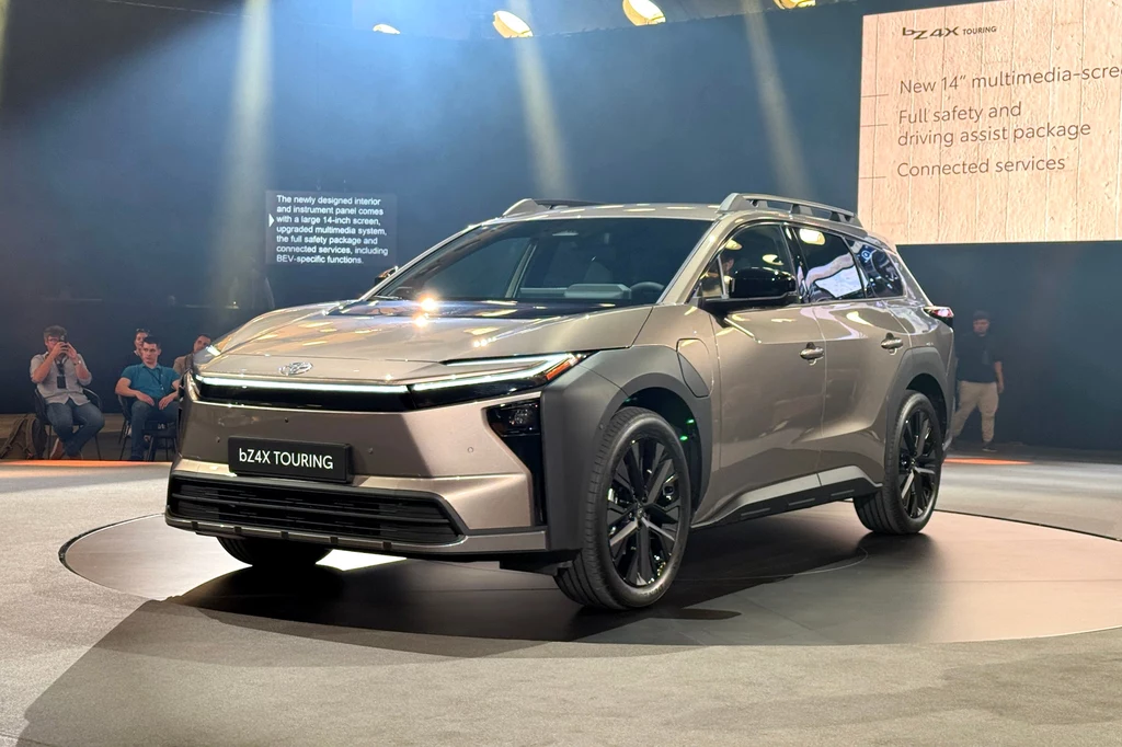 Samochód typu SUV prezentowany na tle sceny wystawowej, otoczony reflektorami. Na przodzie pojazdu widoczne logo producenta oraz oznaczenie modelu bZ4X Touring. W tle kilka osób obserwujących prezentację samochodu.