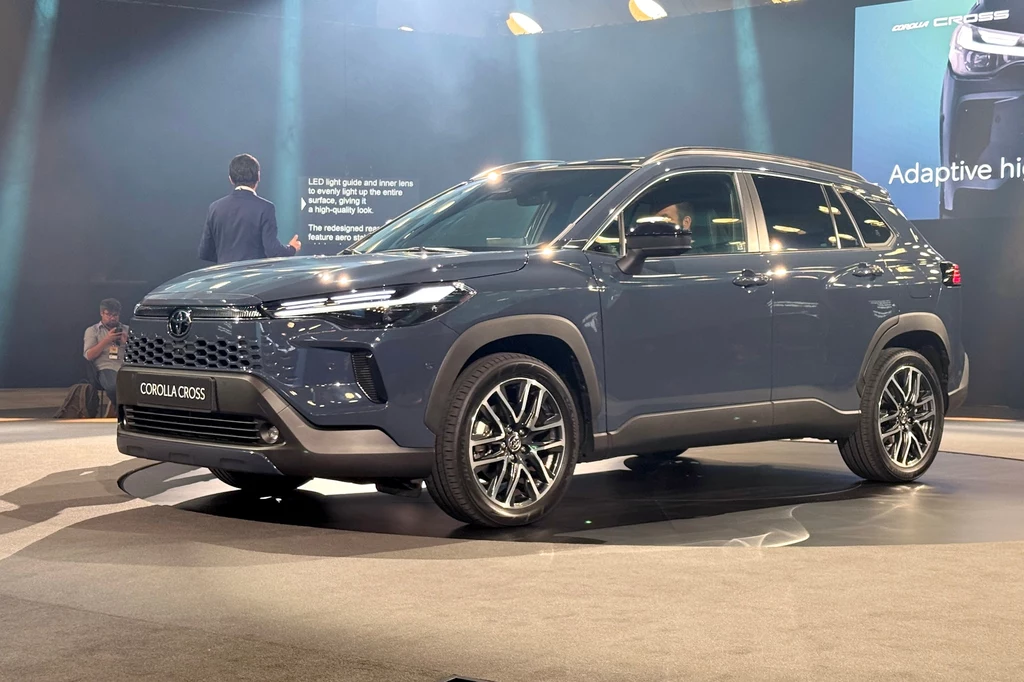 Nowoczesny SUV Corolla Cross w ciemnym kolorze prezentowany na scenie podczas wydarzenia targowego. W tle widoczni są ludzie oraz ekrany wyświetlające informacje o modelu.