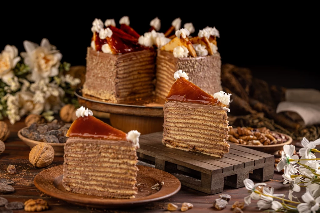 Tort Dobosza to eksplozja smaku - cieniutkie warstwy biszkoptów przekładane kremem czekoladowym, z karmelową polewą Elegancko podane ciasto waflowe z warstwami nadzienia czekoladowego, ozdobione śmietanką i karmelem, na drewnianej desce, w otoczeniu kwiatów i orzechów, co tworzy rustykalną atmosferę.