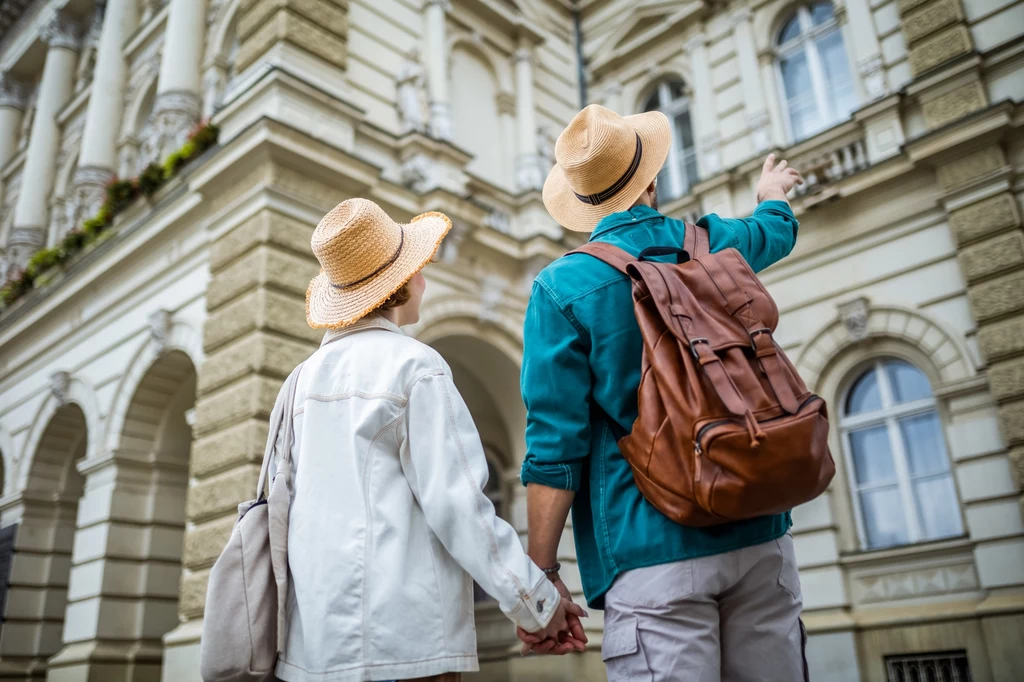 Program "CopenPay" opłaca się zarówno władzom miasta jak i turystom Para turystów w kapeluszach słomkowych, z plecakami, stoi przed imponującym budynkiem w klasycznym stylu, patrząc w górę. Mężczyzna wskazuje w stronę okna na wyższym piętrze, co może świadczyć o ich zainteresowaniu detalami architektonicznymi.