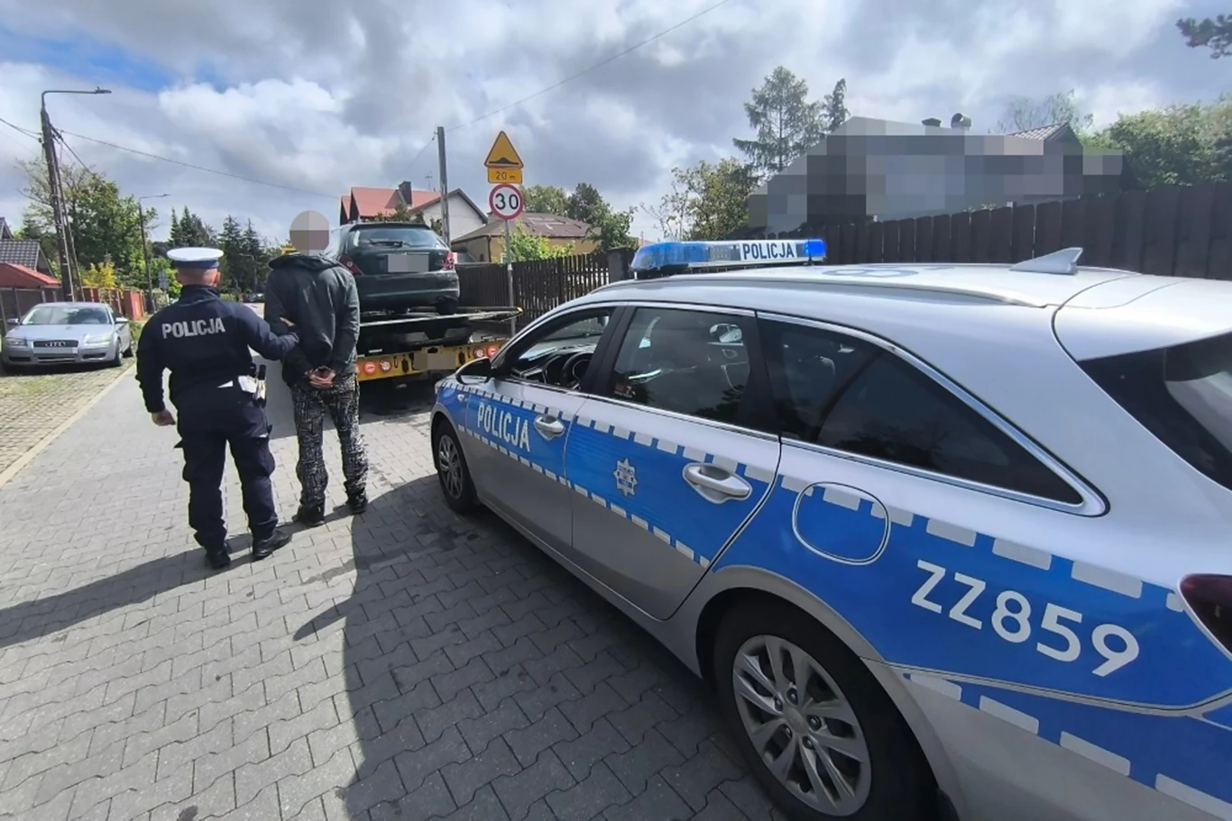 Funkcjonariusze miejscowej drogówki natknęli się na nietypowy widok. Policja zatrzymuje osobę na tle radiowozu, a na lawecie w tle znajduje się pojazd. Na ulicy widoczne są znaki drogowe oraz inne samochody zaparkowane wzdłuż drogi.