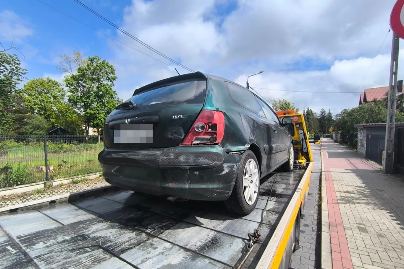 Pojazd miał uszkodzoną tylną lampę oraz całkowicie zniszczony przód Uszkodzony samochód stoi na lawecie, na tle widać zadrzewioną ulicę z domami po jednej stronie. Pojazd został uszkodzony z tyłu, co wskazuje na kolizję. Niebo jest częściowo pochmurne.