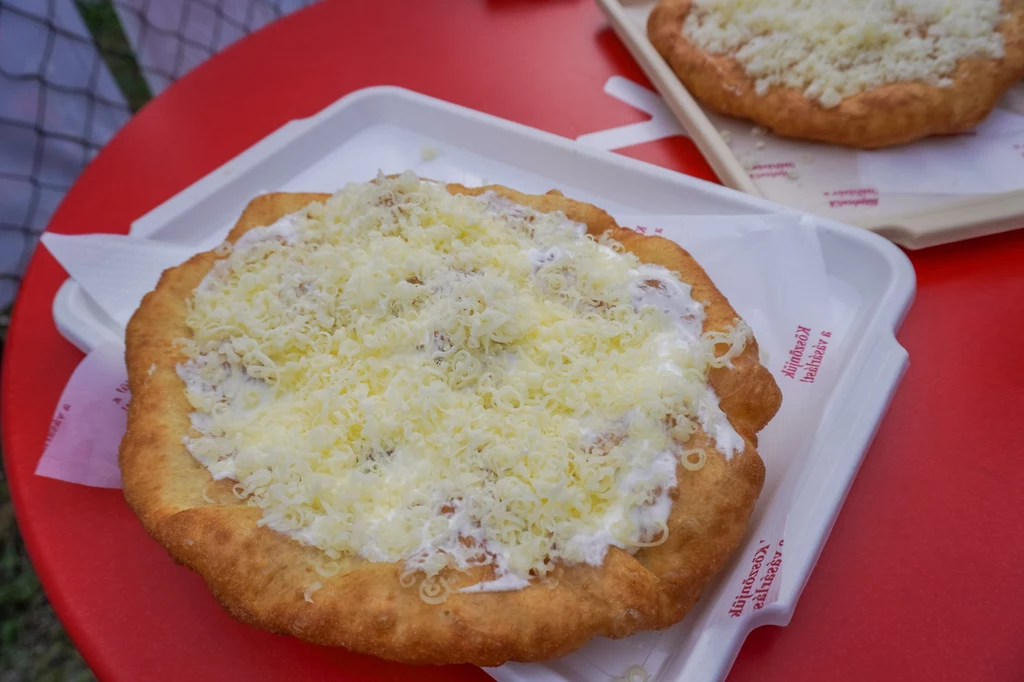 Langosze to węgierski street food, który możesz odtworzyć sama w domu! Langosz na plastikowej tacy, pokryty serem i śmietaną. W tle widoczny drugi langosz na czerwonym stoliku.