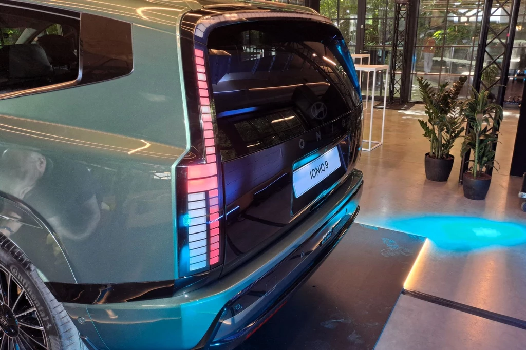 Hyundai Ioniq 9 dostępny będzie w trzech wariantach napędu. Niezależnie od wyboru, za magazynowanie energii odpowiadać będzie akumulator o pojemności 110,3 kWh. Samochód elektryczny z otwartym bagażnikiem, widoczne charakterystyczne tylne światła LED. Wnętrze jest dobrze oświetlone, a otoczenie to nowoczesne wnętrze z roślinami w doniczkach i przeszkleniami.