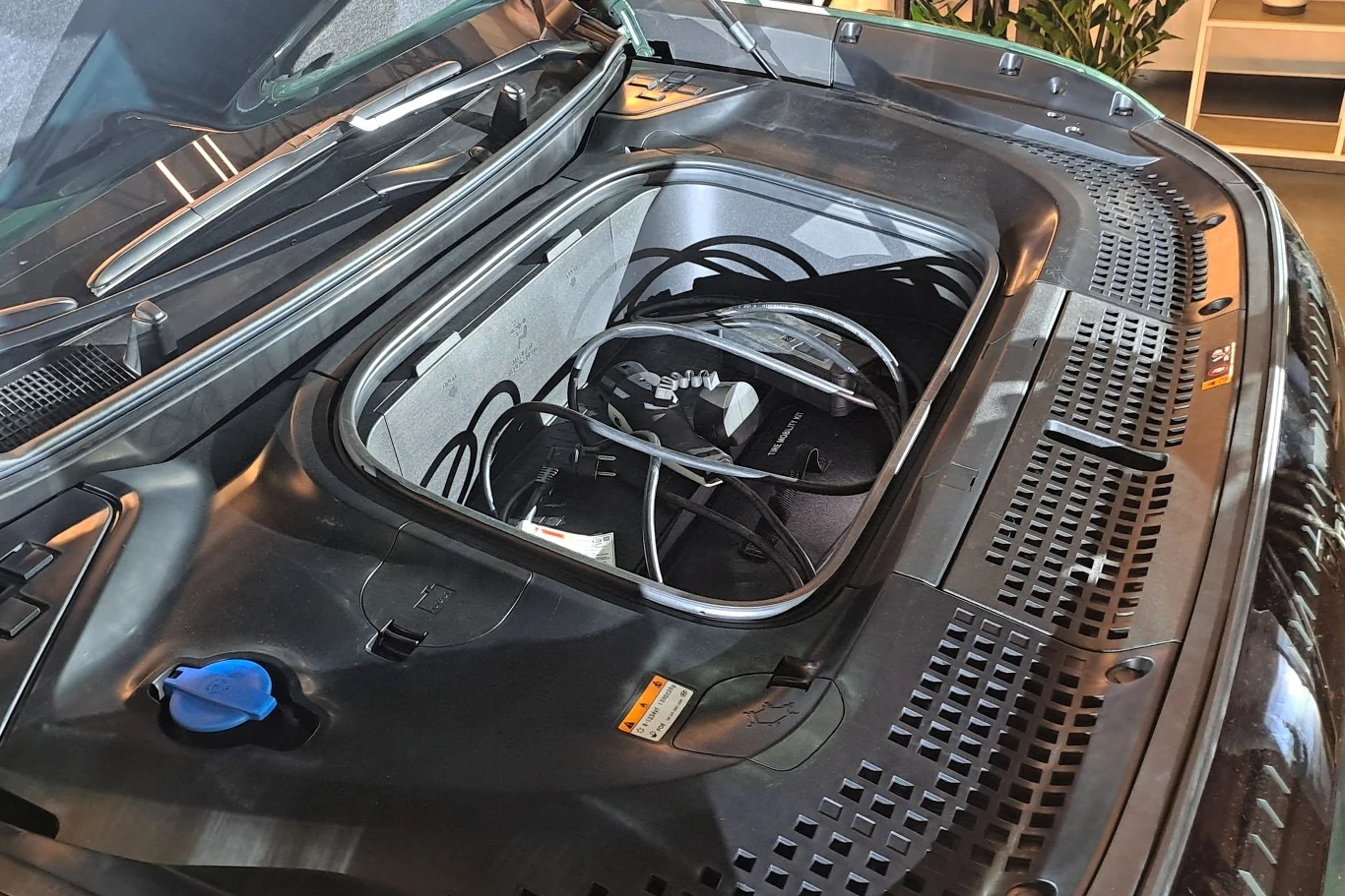 Przednia część elektrycznego samochodu z otwartą przestrzenią bagażową pod maską, zawierającą przewody i akcesoria elektroniczne. Pokrywa w czarnym kolorze z niebieskim korkiem od płynu chłodzącego po lewej stronie.