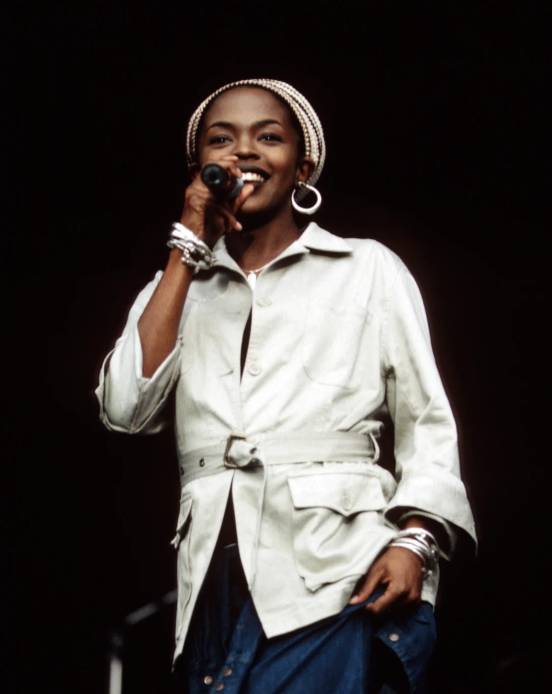 Lauryn Hill Osoba śpiewająca na scenie, trzymająca mikrofon, ubrana w jasną kurtkę i niebieskie spodnie. Uśmiecha się do publiczności, ma na sobie charakterystyczne kolczyki i bransoletki.