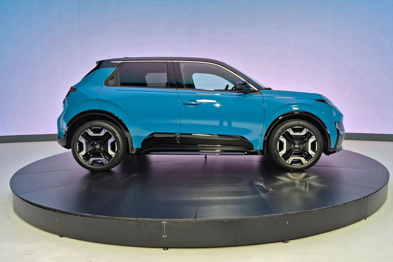 Nowoczesny, niebieski SUV na obrotowym podeście, z widoczną boczną sylwetką i dużymi, charakterystycznymi felgami. Tło jest jednolite, lekko fioletowe, co podkreśla wygląd pojazdu.