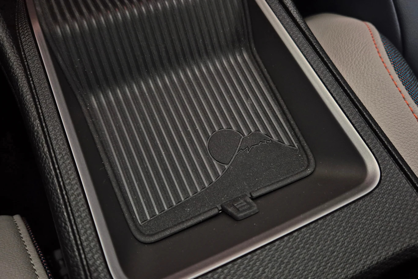 Schowek w samochodzie z żebrowanymi wykończeniami, w którym znajduje się symboliczny wzór góry. Ściany schowka wykonane są z czarnego materiału, a na jego dnie znajduje się subtelne logo. Przestrzeń jest czysta i zorganizowana.