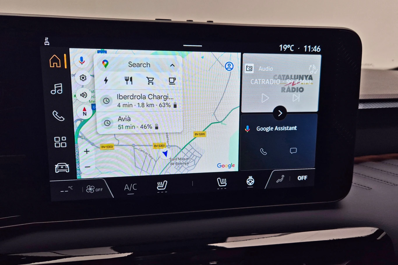Ekran systemu multimedialnego w samochodzie, ukazujący mapę nawigacyjną z zaznaczonymi stacjami ładowania oraz interfejs audio z radiem i asystentem Google.