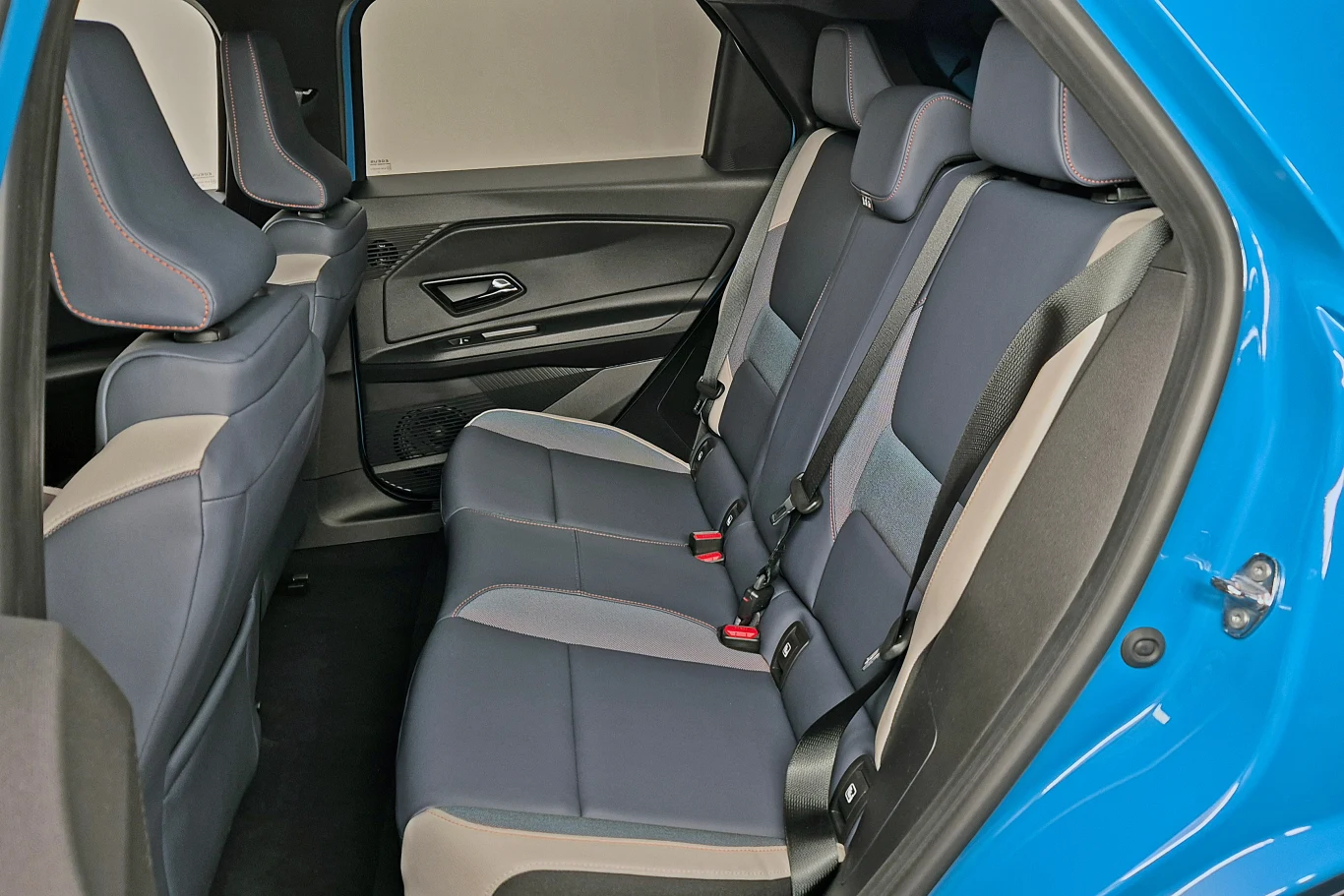 Wnętrze tylnej części samochodu z widocznymi siedzeniami w ciemnym kolorze, dopasowanymi zagłówkami i pasami bezpieczeństwa. Drzwi boczne otwarte, ukazujące detale wnętrza.