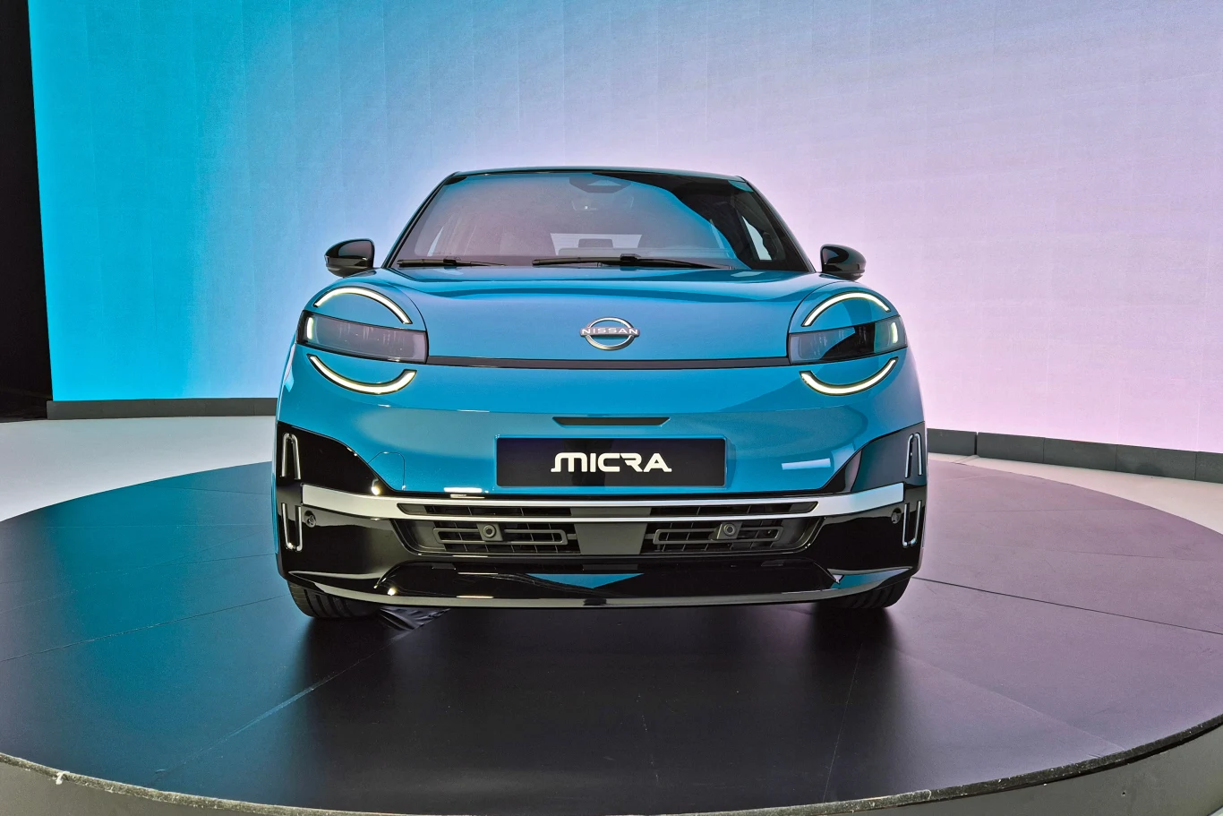 Samochód Nissan Micra o nowoczesnym designie, wyeksponowany na obrotowej platformie. Przód pojazdu charakteryzuje się futurystycznymi reflektorami i logo marki na masce. Tło utrzymane w jasnych kolorach, co podkreśla estetykę auta.