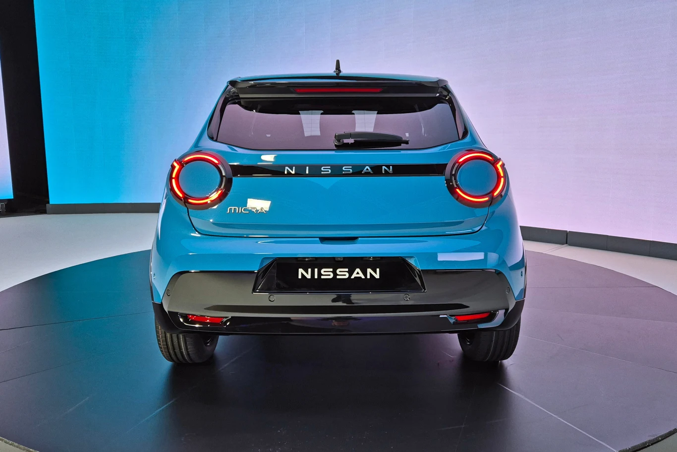 Tylna część niebieskiego samochodu Nissan Micra na obrotowej platformie, z charakterystycznymi okrągłymi tylnymi światłami oraz logo marki.