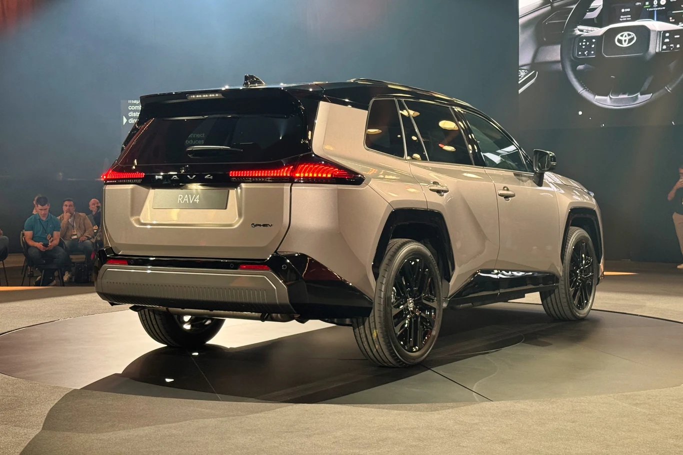Nowoczesny samochód terenowy Toyota RAV4 prezentowany na tle sceny oświetlonej reflektorami, z widocznymi eleganckimi liniami nadwozia i matowym wykończeniem. Grupa osób siedzących z boku sali, obserwująca prezentację.
