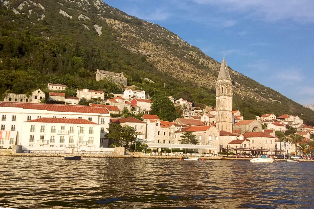 Rejs ma jeszcze jedną zaletę. Pozwala spojrzeć na miasteczko z kolejnej perspektywy Widok na malownicze miasteczko położone nad wodą, w otoczeniu gór. Na pierwszym planie widać zabytkowe budynki z charakterystycznymi czerwonymi dachami, a centralnym punktem jest wysoka kamienna wieża kościoła. Spokojna tafla wody odbija delikatne prom...