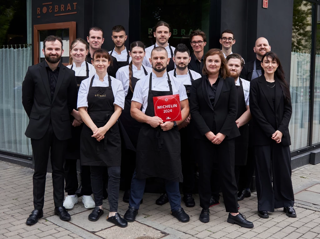 Zespół restauracji Rozbrat Grupa pracowników restauracji Rozbrat z uśmiechem pozuje do zdjęcia przed wejściem do lokalu. W centrum stoi mężczyzna z czerwoną książką Michelin 2024, co świadczy o wyróżnieniu lokalu przez przewodnik Michelin.