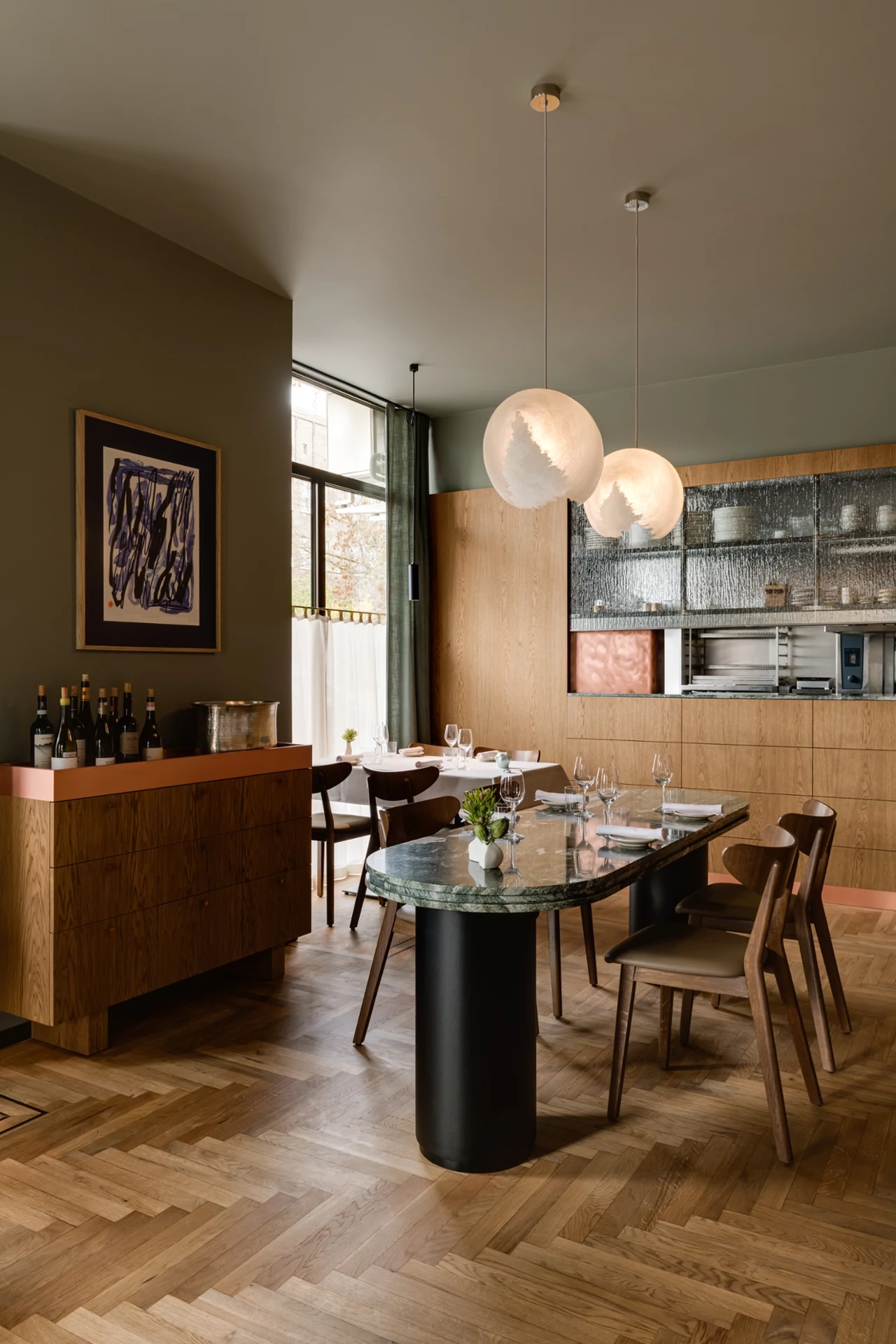 Wnętrze restauracji Elegancka jadalnia z nowoczesnym designem, czarnym owalnym stołem i krzesłami z drewna. Nad stołem wiszą stylowe lampy, a w tle widoczny jest barek z winami oraz zabudowa kuchenna. Duże okno wpuszcza naturalne światło.