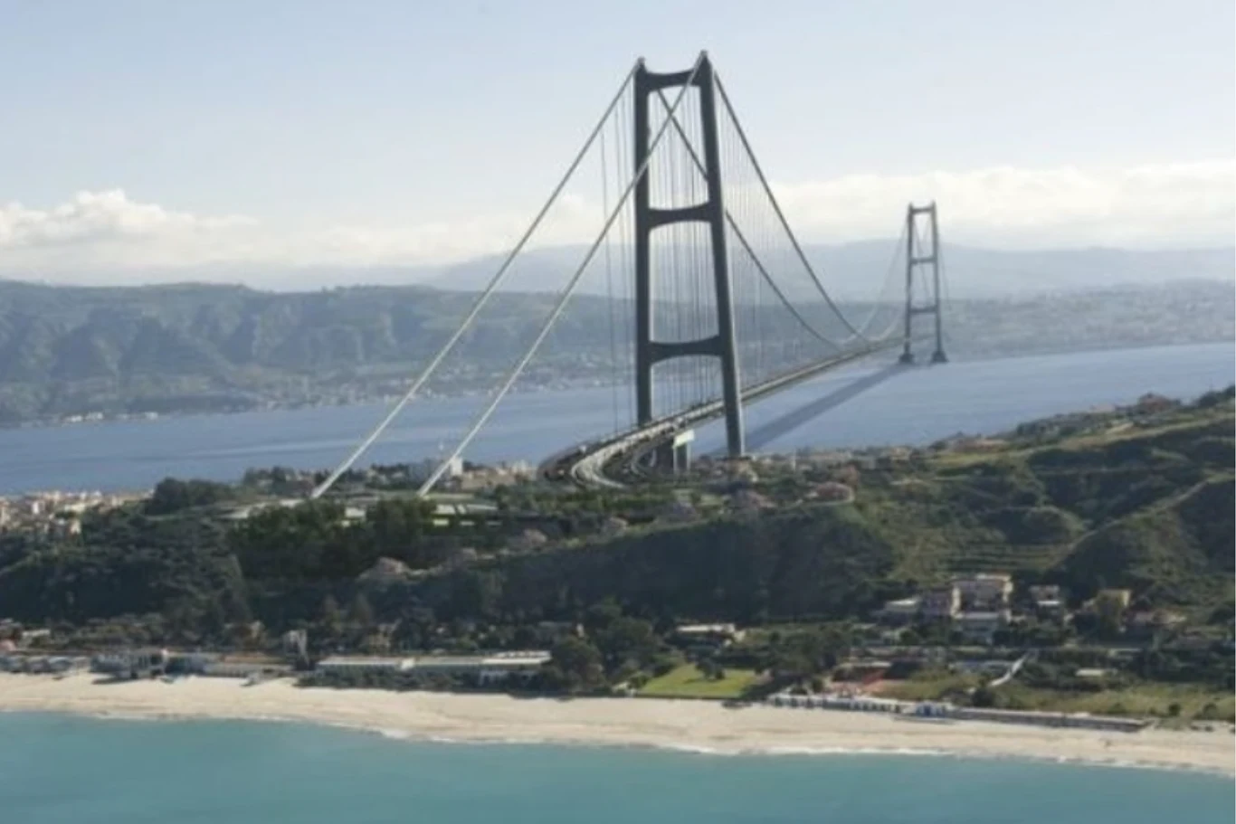 Most wiszący nad cieśniną otaczającą zielone wzgórza, z widocznym wybrzeżem i plażą. W oddali można dostrzec rozległe pasma górskie i otwarte morze.