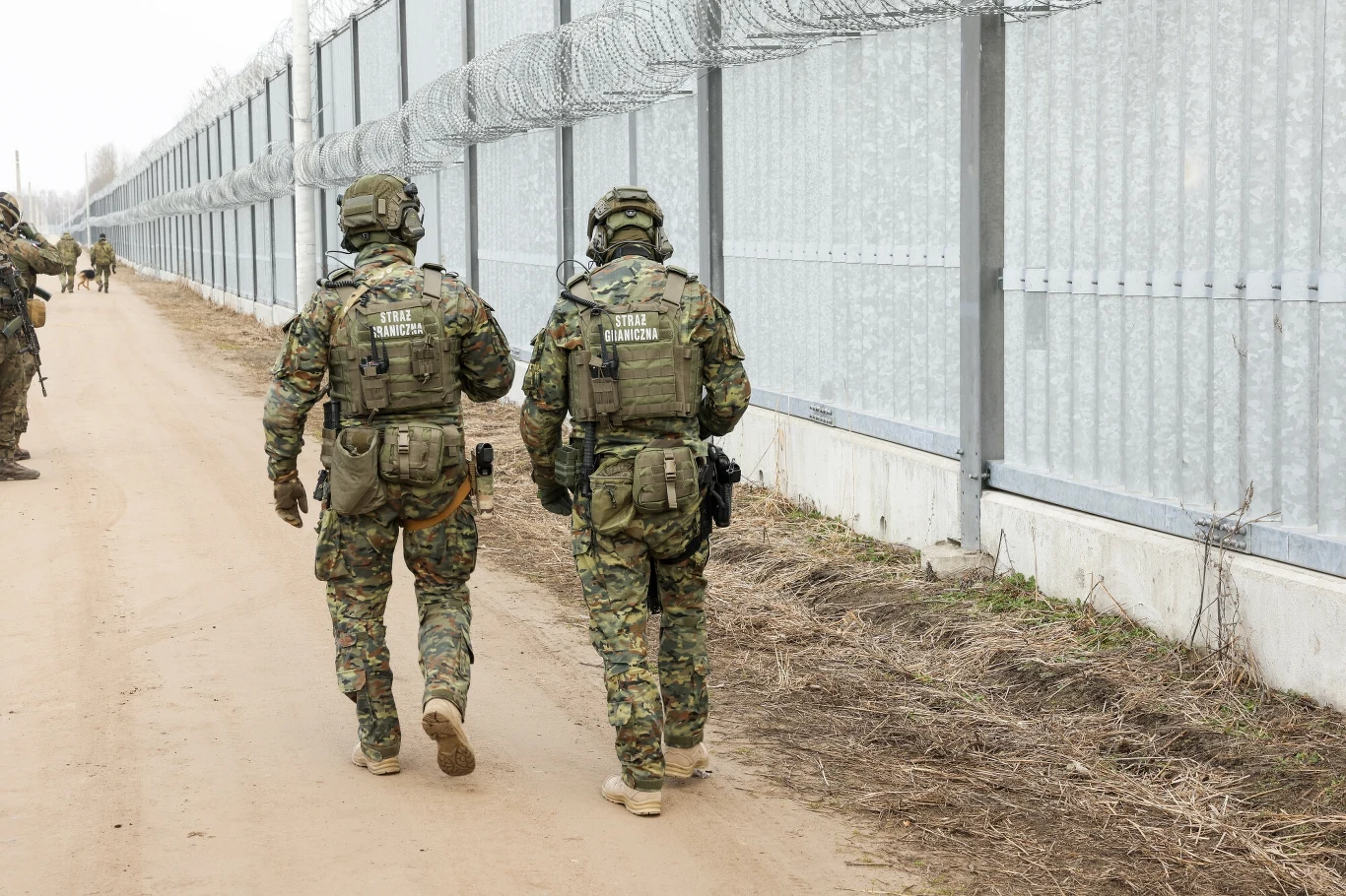 Granica polsko-białoruska Dwóch żołnierzy w pełnym umundurowaniu taktycznym patroluje drogę wzdłuż wysokiego ogrodzenia zabezpieczonego drutem kolczastym, na ziemi widoczna jest sucha, ubita trawa i ziemia, w tle kolejni żołnierze.