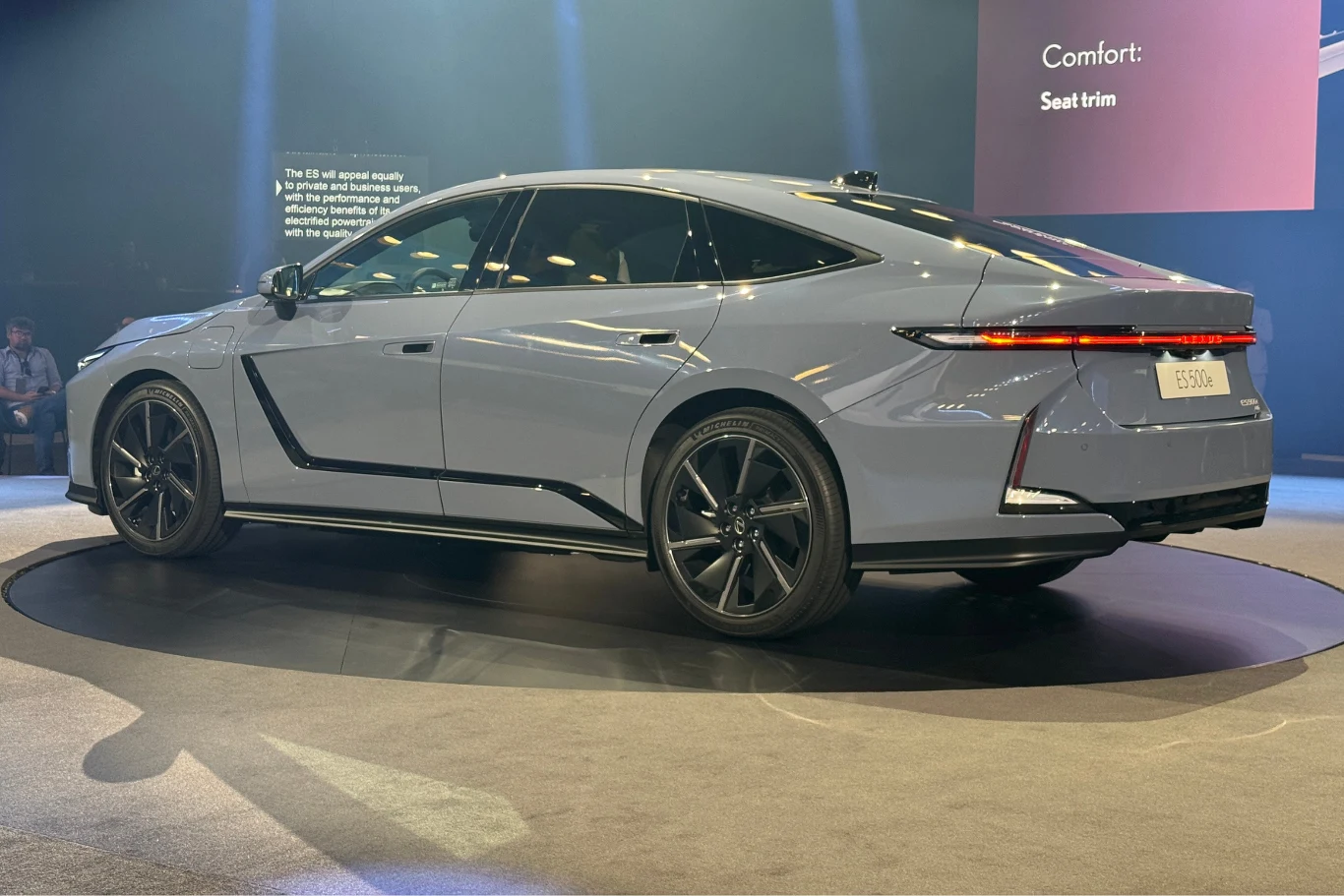 Lexus ES jest dłuższy względem poprzednika o 16,5 cm. Nowoczesny srebrny samochód osobowy typu sedan ustawiony na podium podczas prezentacji, widoczne światła LED z tyłu, eleganckie linie nadwozia i sportowe felgi, w tle oświetlenie sceniczne oraz fragment ekranu prezentacyjnego.