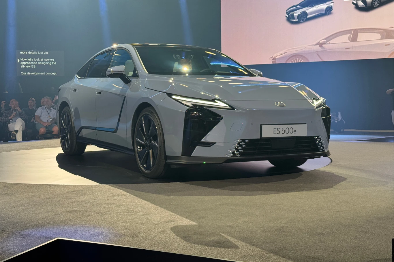 Tak wygląda ósma generacja Lexusa ES. Jesienią poznamy polskie ceny. Nowoczesny elektryczny samochód Lexus ES 500e w jasnoszarym kolorze zaprezentowany na scenie podczas oficjalnej premiery, światła reflektorów skierowane na auto, za nim widoczne grafiki i fragmenty publiczności w tle.