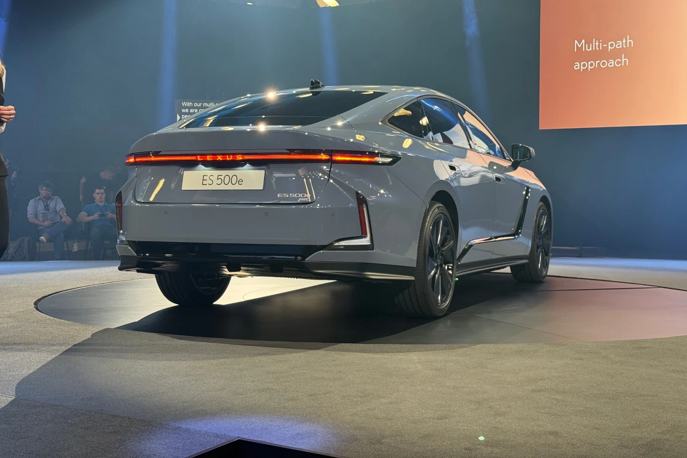 Motyw litery "L" pojawia się zarówno w przednich, jak i tylnych lampach. Srebrny samochód elektryczny marki Exeed ES 500e prezentowany na scenie podczas wydarzenia motoryzacyjnego, otoczony przez publiczność i światła prezentacyjne, w tle widoczny napis „Multi-path approach”.
