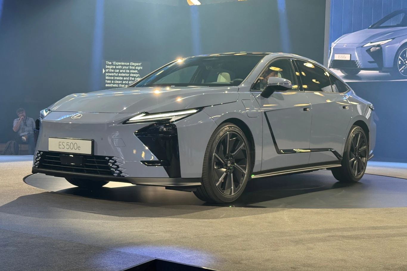 Minimalistyczny design Lexusa ES ósmej generacji może się podobać. Nowoczesny sedan elektryczny w srebrnym kolorze stoi na ekspozycji podczas targów motoryzacyjnych, w tle rozmyte światła sceniczne i wyświetlacz z grafiką pojazdu.